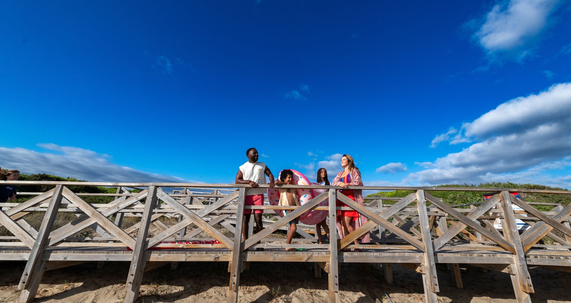 Familie med børn står på bro på stranden og ser ud over vandet